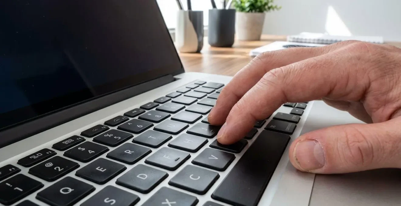 Detalle de una mano escribiendo en el teclado de un portátil moderno con pantalla difuminada mostrando gráficos en segundo plano